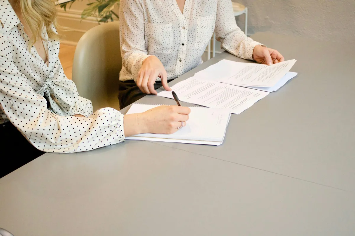 Two Women Working Together To Get Work Delegated via Paperwork sitting at a desk with a pen in their hands.